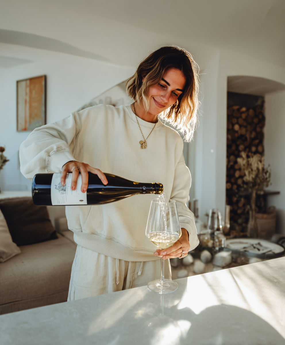 A woman pouring a glass of white wine in a sunlit kitchen